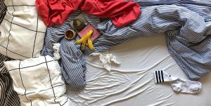 Der Ausschnitt eines Bettes von oben: Darauf blau gestreifte zerknautschte Bettwäsche, ein Kaffeebecher, eine Bananenschale und ein rotes Tuch.