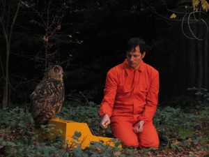 Felix Kubin in rotem Overall sitzt im Wald, vor ihm ein gelber Kasten, worauf eine Eule sitzt.
