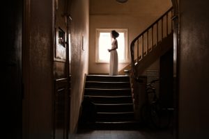 Eine Schwarze Frau steht weit hinten im Bild in einem langen weißen Kleid auf einem Treppenabsatz vor einem Fenster im Gegenlicht. Eine:r sieht sie von der Seite, ihre Arme sind gebeugt.