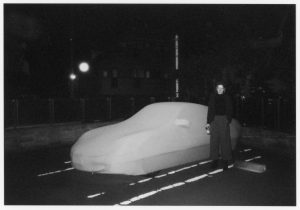 Schwarz-weiß Foto bei Nacht mit einer Person, die mit einer Flasche in der Hand vor einem mit einem weißen Bezug ummantelten Auto steht.