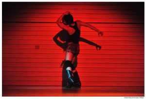 Tänzerin Jolson hält ihre eine Hand auf den Kopf und streckt die andere nach hinten. Sie befindet sich vor einer rot beleuchteten Wand.