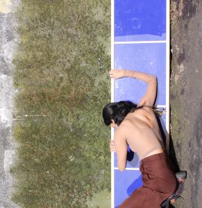 Eine Performerin liegt auf einem blauen Streifen mit weißer Umrahmung, der sich wiederum auf einem mit Moos bewachsenen grauen Boden befindet.