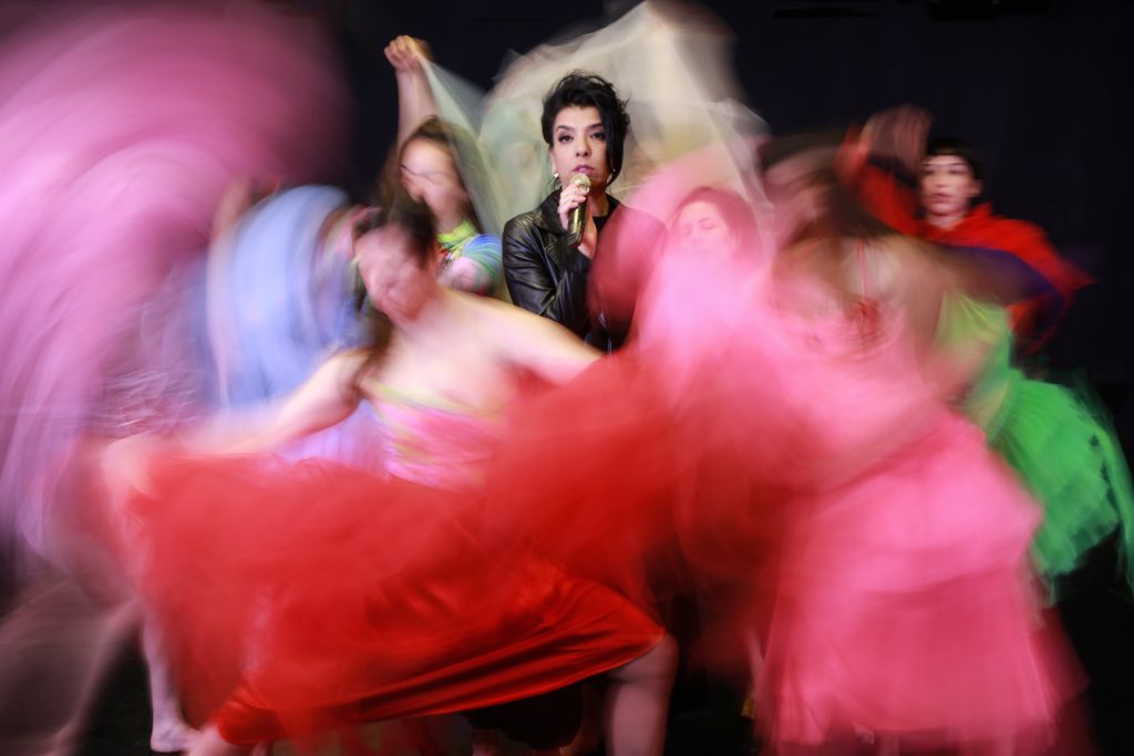 Auf dem Foto steht die Sängerin Dikla, gekleidet in schwarzer Lederjacke, zwischen den Tänzerinnen in bunten Kleidern. Während Dikla in die Kamera guckt und gut erkennbar ist, sind die Tänzerinnen um sie herum mitten in Bewegung und ihre blauen, roten, grünen und pinken langen Kleider sind aufgrund der Bewegung nur unscharf zu erkennen.